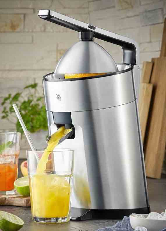 Elektrische Zitruspresse füllt ein Glas mit frischem Orangensaft, daneben halbierte Zitrusfrüchte auf der Arbeitsplatte