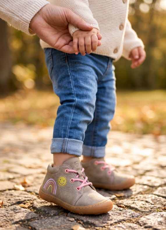 Kind in Jeans und bunten Schnürschuhen mit Regenbogenmotiv läuft draußen und hält die Hand einer erwachsenen Person