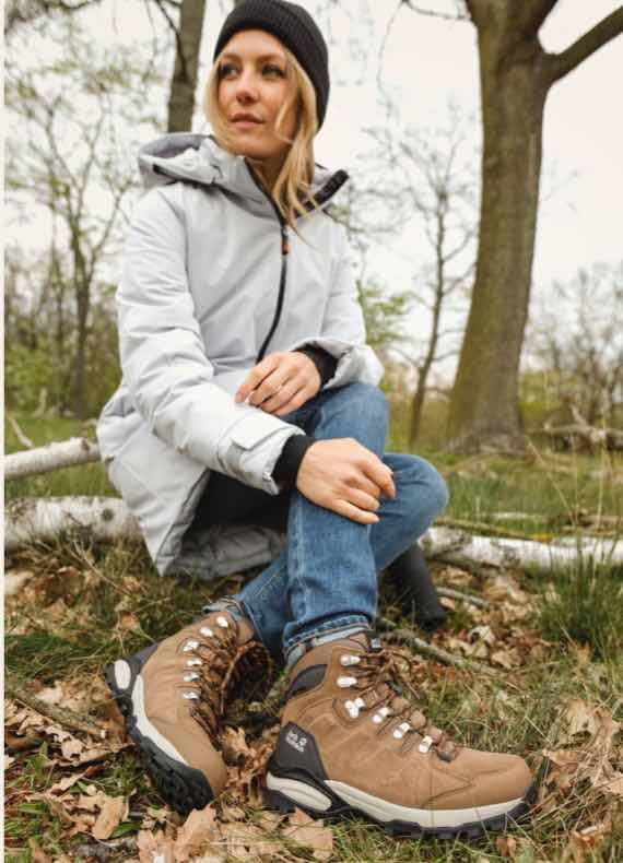 Frau im Parka und Mütze sitzt im Wald auf dem Boden und präsentiert braune Wanderstiefel am Fuß