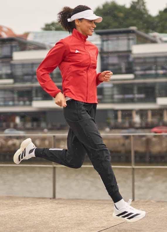 Frau joggt am Wasser, trägt rote Laufjacke, schwarze Jogginghose, weiße Kappe und Sportschuhe mit drei Streifen