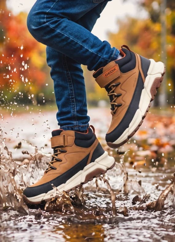 Kind springt mit braunen Sneakers in Pfütze, Wasser spritzt hoch, herbstliche Bäume im Hintergrund