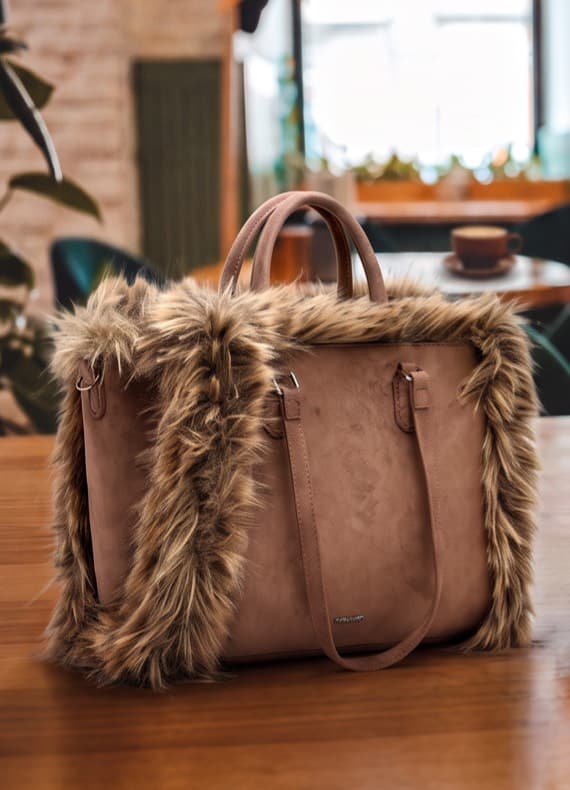 Große beigefarbene Handtasche mit breitem, pelzbesetztem Rand steht auf einem Holztisch in einem Café-Hintergrund