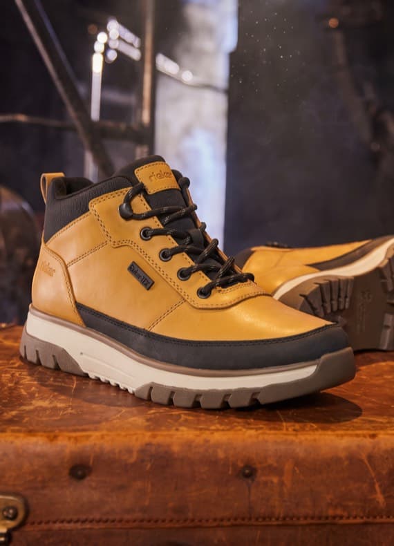 Gelb-schwarzer knöchelhoher Schnürschuh mit Profilsohle steht auf braunem Koffer, zweiter Schuh im Hintergrund