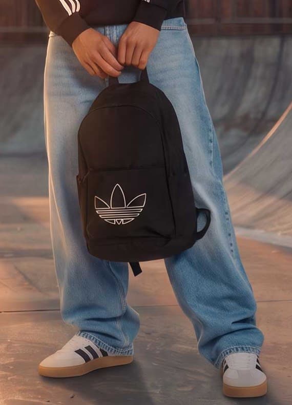 Person im Skatepark hält schwarzen Rucksack mit großem Adidas-Trefoil-Logo, trägt weite Jeans und Sneakers
