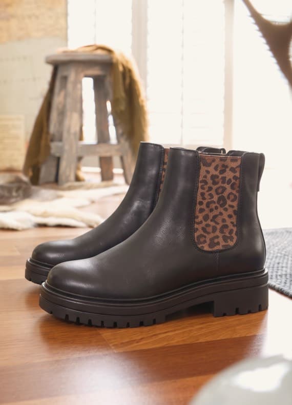 Schwarze Chelsea-Boots mit seitlichem Gummieinsatz im Leopardenmuster auf Holzboden in wohnlicher Umgebung