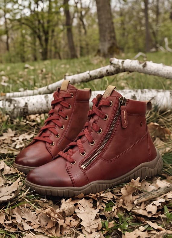 Rote knöchelhohe Schnürstiefel mit seitlichem Reißverschluss stehen auf Herbstlaub im Wald vor hellen Birkenstämmen