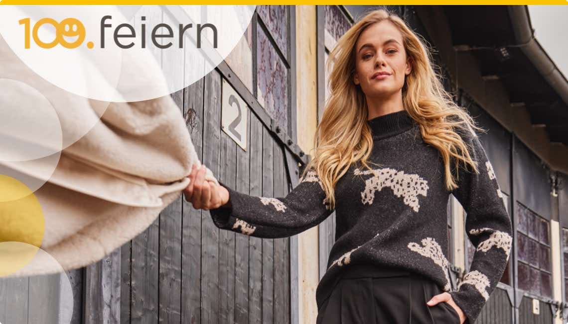 100. feiern – Frau in schwarzem Pullover mit Tiermuster hält eine beige Decke vor einem Gebäude mit der Nummer 2