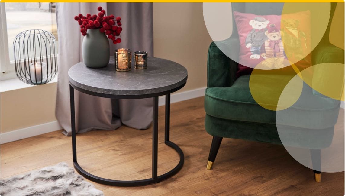 Runder Beistelltisch mit Kerzen und Vase, daneben grüner Sessel mit Kissen, auf dem zwei Teddybären abgebildet sind