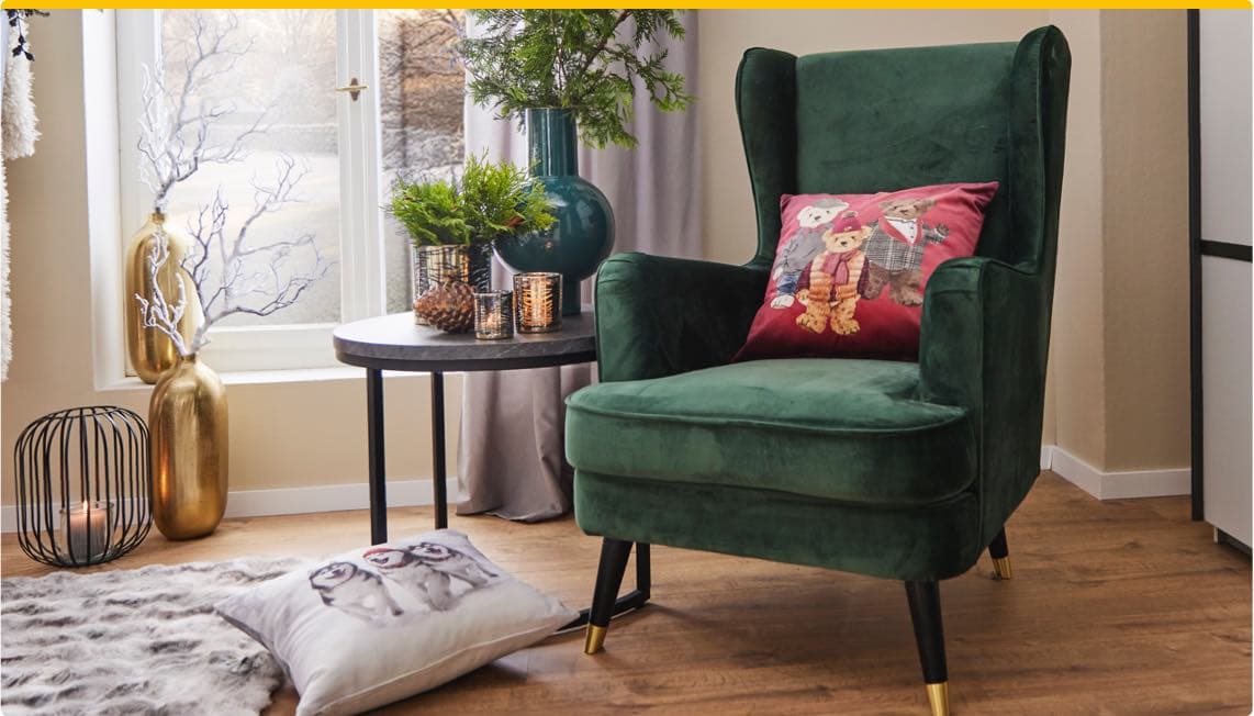 Gemütliche Ecke mit grünem Sessel, rotem Kissen mit Bärenmotiv, rundem Tisch mit Deko und Kissen mit Hundemotiv auf Teppich