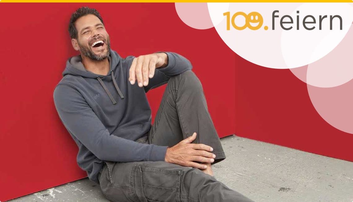 100.feiern - Mann in grauem Kapuzenpullover und dunklen Hosen sitzt entspannt an roter Wand