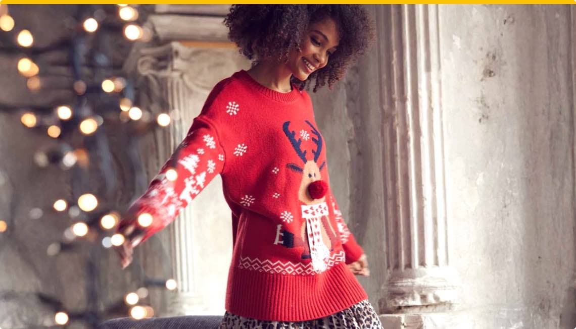 Frau trägt roten Weihnachtspullover mit Rentiermotiv und Schneeflocken, posiert vor einer Wand mit Säulen und Lichterkette im Hintergrund