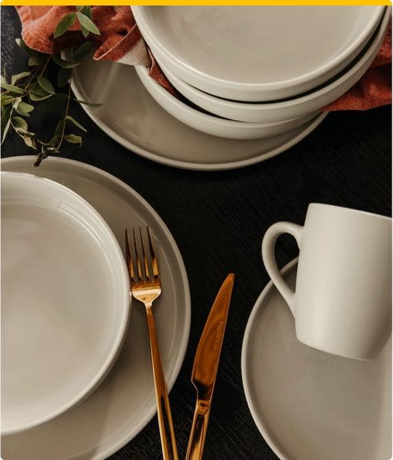 Weißes Geschirrset mit Tellern, Schalen und Tassen, dazu goldfarbene Gabel und Messer, auf schwarzem Tisch arrangiert