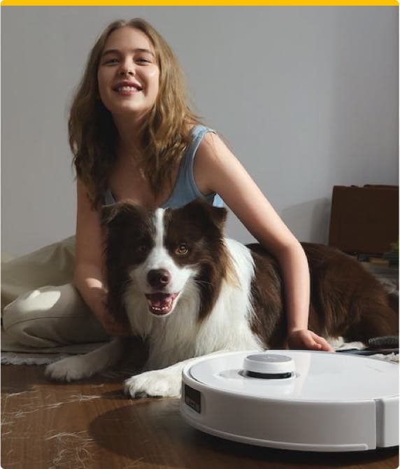 Frau sitzt mit einem braun-weißen Hund auf dem Boden, neben ihnen ein weißer Saugroboter