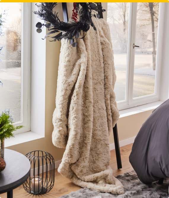 Beige Decke hängt über einen Stuhl neben einem Fenster, im Vordergrund ein schwarzes Drahtkorb mit Kerze.