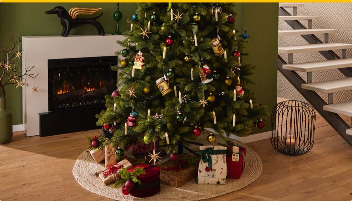 Weihnachtsbaum mit Kerzen und bunten Kugeln, Geschenke darunter, Kaminfeuer und Treppe im Hintergrund.