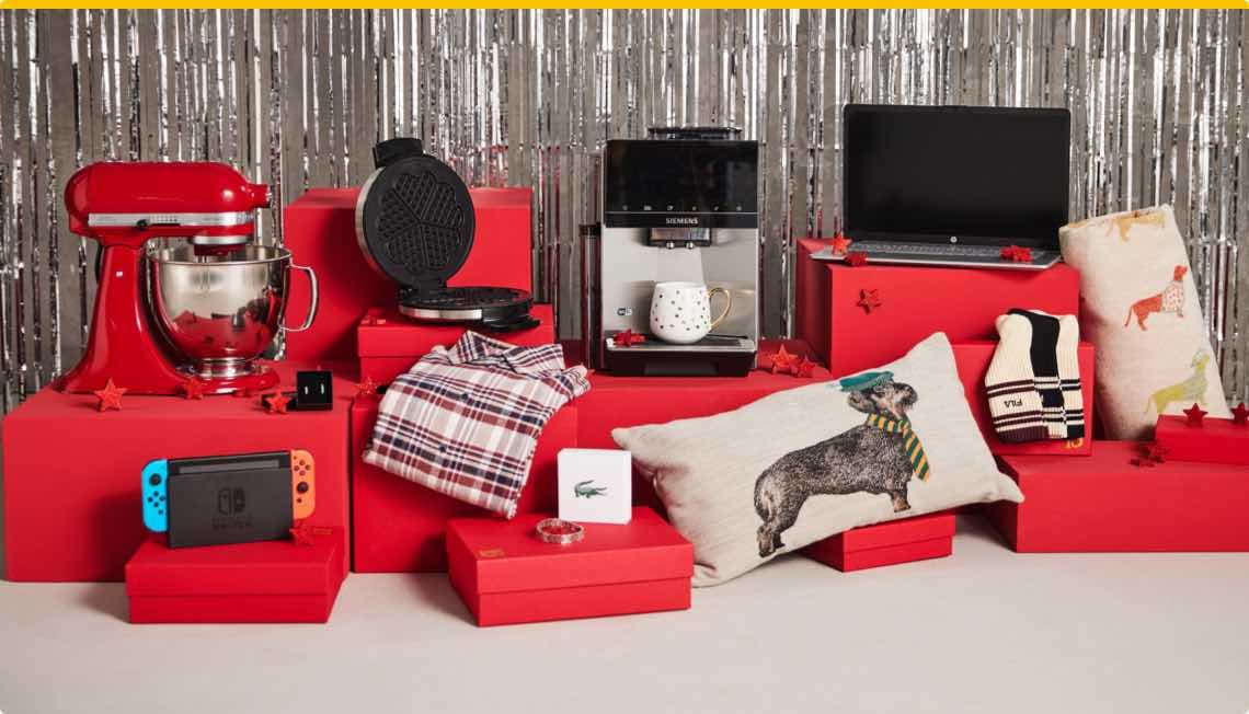 Geschenke auf roten Boxen vor silbernem Hintergrund, darunter Mixer, Waffeleisen, Kaffeemaschine, Laptop, Kissen und Kleidung