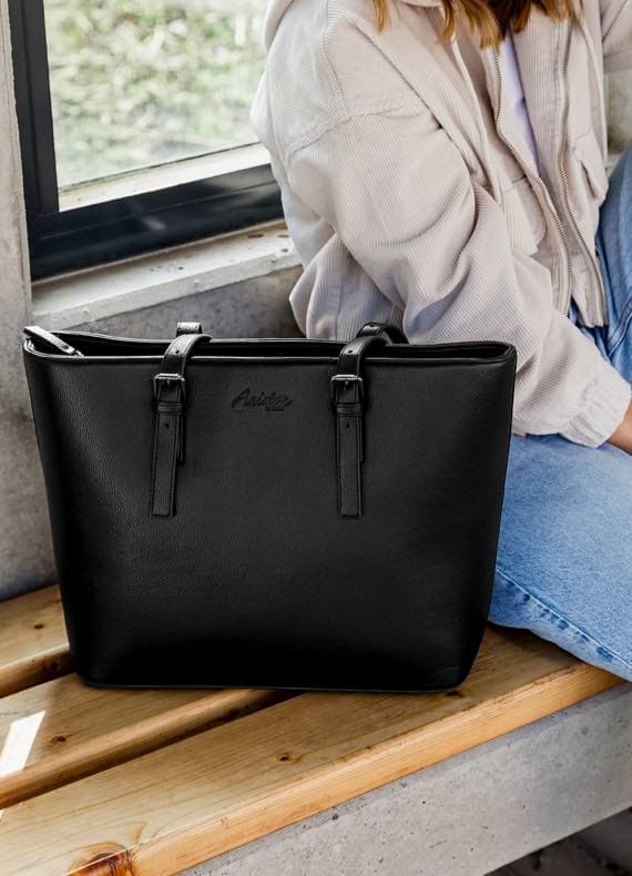 Schwarze Tasche auf Holzbänken, Frau im Hintergrund trägt beige Jacke und blaue Jeans.