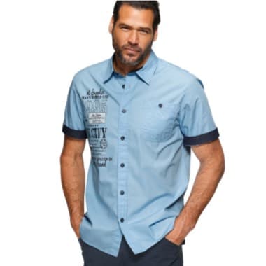 Mann in hellblauem Hemd mit Aufdruck und dunkelblauen Akzenten, Hände in den Hosentaschen.