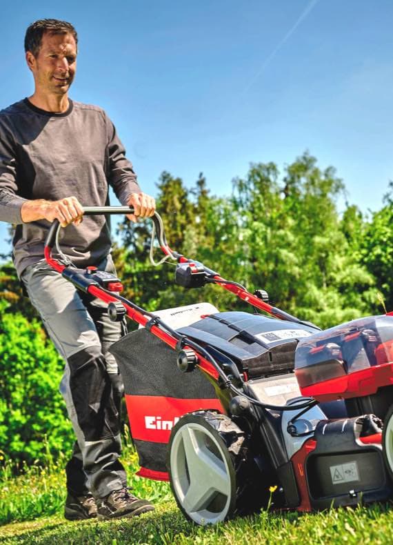 Person schiebt einen Einhell Rasenmäher auf einer grünen Wiese bei sonnigem Wetter.