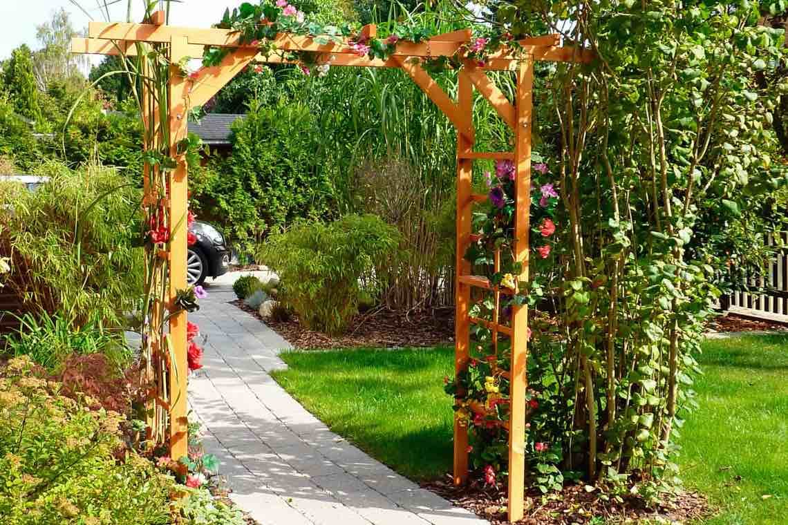 Gartenweg führt durch einen bepflanzten Torbogen mit Rankpflanzen, dahinter Rasen und Sträucher