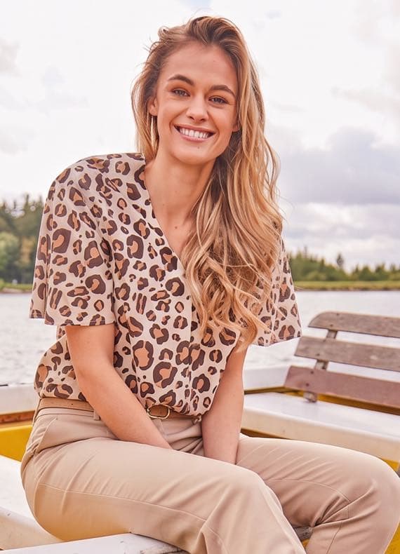 Frau im Leopardenmuster-Shirt und hellen Hosen sitzt auf einer Bank im Boot vor einem See