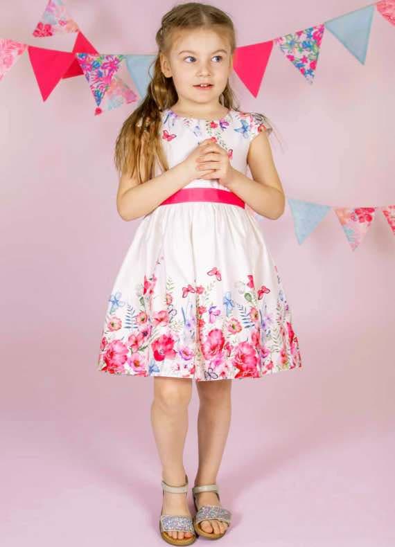 Mädchen im weißen Kleid mit buntem Blumenrand und pinkem Gürtel vor rosa Hintergrund mit Wimpelgirlande