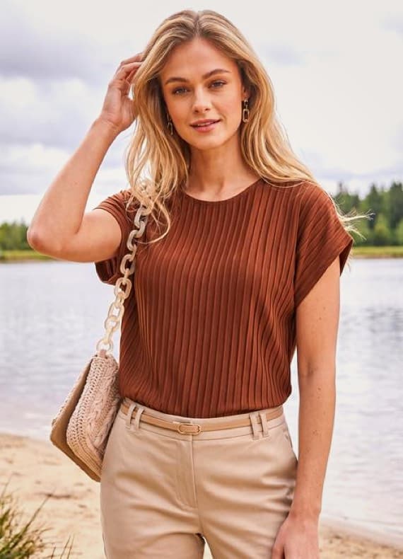 Frau im braunen Faltshirt und hellen Hose steht mit Umhängetasche am Seeufer