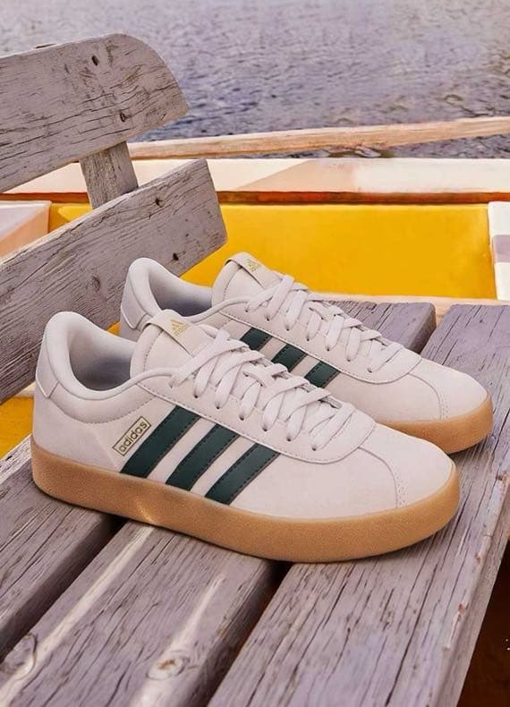 Weiße Adidas Sneaker mit dunklen Streifen liegen paarweise auf verwitterter Holzbank am Wasser