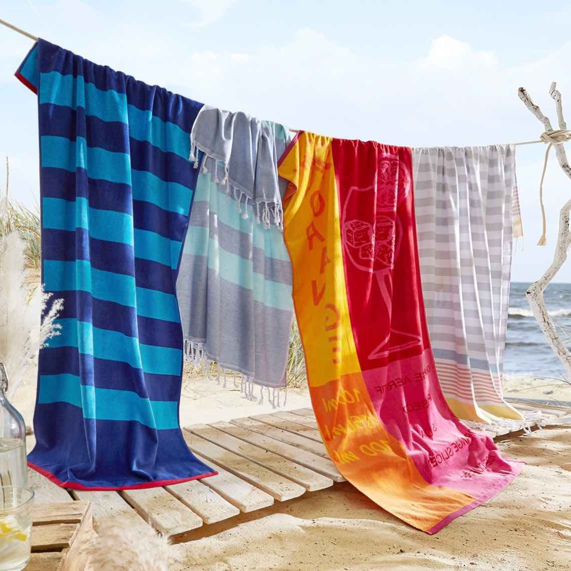 Bunte Strandtücher in Streifen und mit Motiv hängen an einer Leine über Holzsteg am Sandstrand mit Meer im Hintergrund