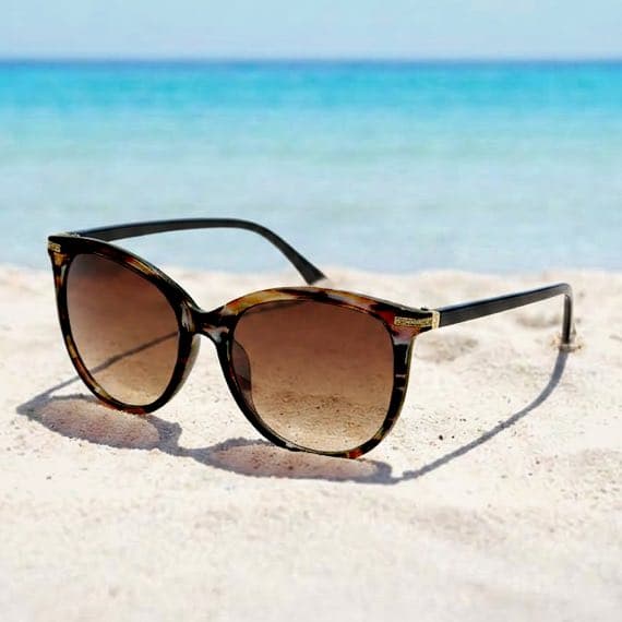 Sonnenbrille mit dunklen Gläsern liegt im hellen Sandstrand vor ruhigem, blauem Meer