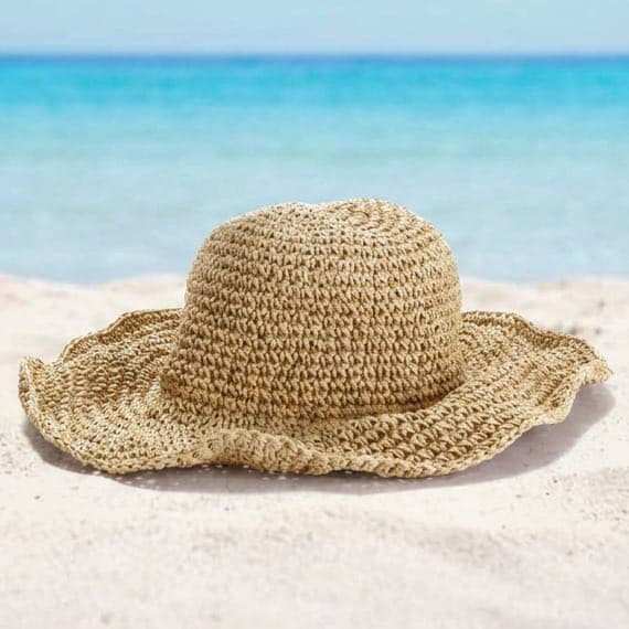 Beiger Sommerhut mit breiter Krempe liegt im Sand vor türkisblauem Meer und hellem Himmel