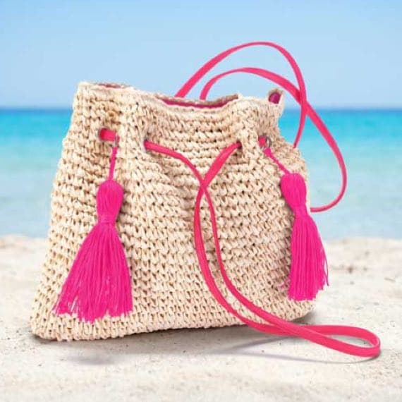 Beige Strandtasche mit langen pinken Quasten und Schulterriemen liegt im Sand vor türkisblauem Meer