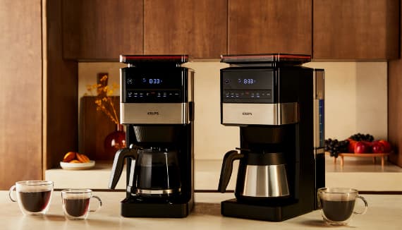 Zwei Krups-Kaffeemaschinen nebeneinander auf Küchenzeile, mit Glaskanne, Thermokanne und zwei gefüllten Tassen Kaffee