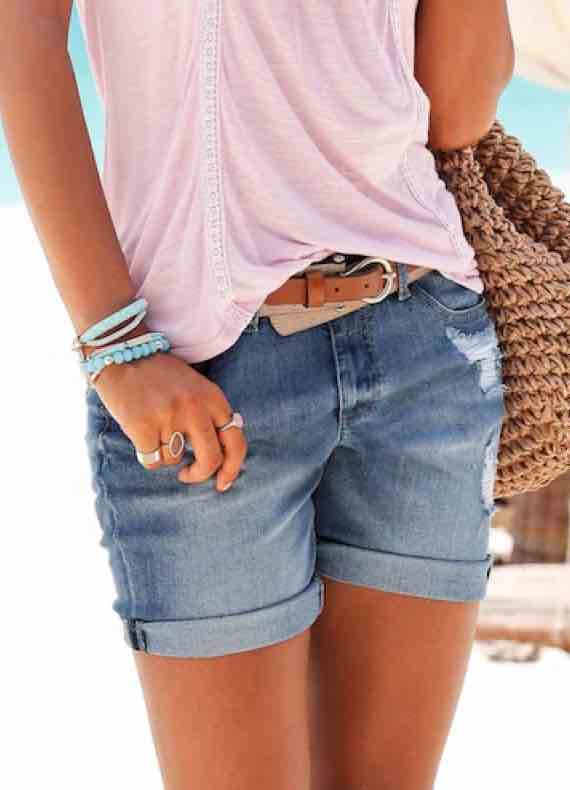 Frau in Jeansshorts und rosa Shirt, mit Gürtel, Armbändern und Flechttasche am Strand