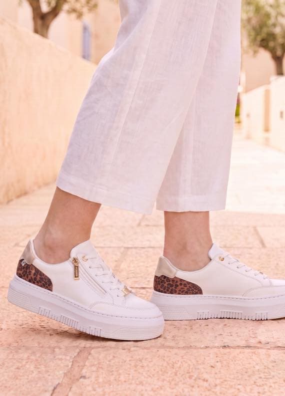 Person in weißen, weiten Hosen trägt weiße Sneaker mit seitlichem Zipper und Leopardenmuster auf gepflastertem Weg