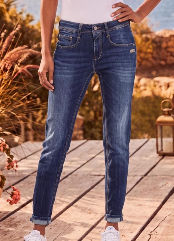 Frau in blauer Jeans mit umgeschlagenen Säumen steht auf Holzterrasse, weißes Top und Sneakers, Pflanzen im Hintergrund