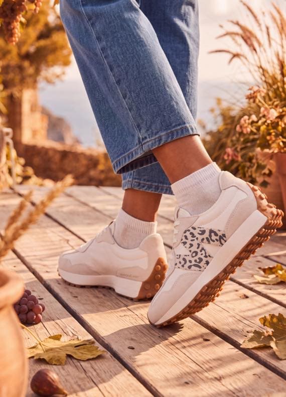 Person in Jeans zeigt beige Sneaker mit gemustertem Einsatz, steht auf Holzterrasse mit Herbstlaub und Trauben