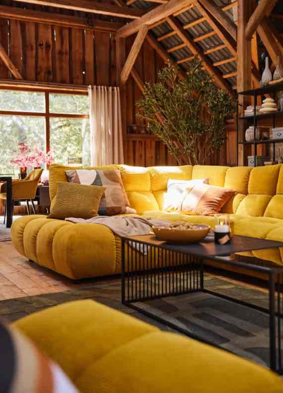 Großes gelbes Ecksofa mit vielen Kissen in rustikalem Wohnzimmer mit Holzwänden, Couchtisch und großem Fenster
