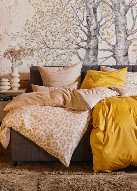 Bett mit gemusterter und senfgelber Bettdecke, vielen Kissen und Wandbild mit Bäumen im Hintergrund