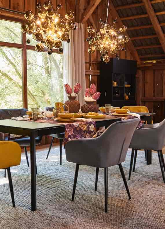 Esstisch mit buntem Geschirr und Blumenläufer, umgeben von grauen und gelben Stühlen unter zwei leuchtenden Kronleuchtern