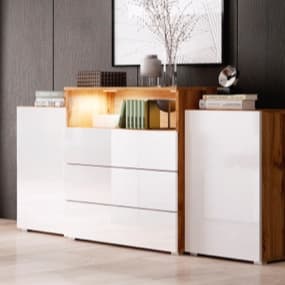 Ein elegantes Sideboard mit einer Kombination aus glänzend weißen und holzfarbenen Elementen. Die Schubladen sind modern gestaltet, und in der Mitte leuchtet eine kleine Lampe, die einige Bücher und Dekorationsobjekte hervorhebt.