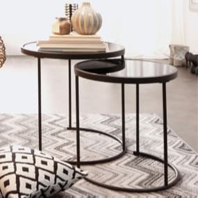 Zwei stilvolle, runde Beistelltische mit schwarzem Glas und einem minimalistischen Metallgestell. Der größere Tisch mit dekorativen Objekten, der kleinere Tisch ist darunter geschoben. Der Teppich darunter hat ein modernes, grafisches Muster.