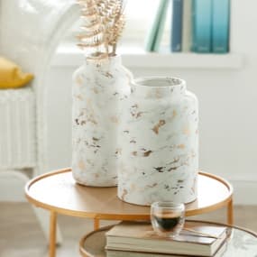 Zwei elegante Vasen mit einem marmorierten Design in Weiß und Gold stehen auf einem runden, kleinen Tisch. Eine der Vasen ist mit dekorativen Zweigen gefüllt. Daneben stehen ein kleines Glas und einige Bücher, was eine einladende Atmosphäre schafft.