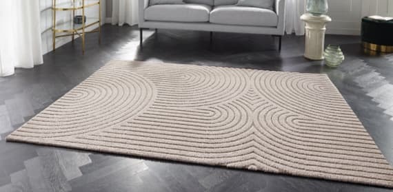 Ein stilvoller Teppich mit einem modernen, Wellen-Muster in sanften Beigetönen liegt auf einem dunklen Holzboden. Im Hintergrund steht ein elegantes Sofa, und neben dem Teppich sind dekorative Möbelstücke platziert.