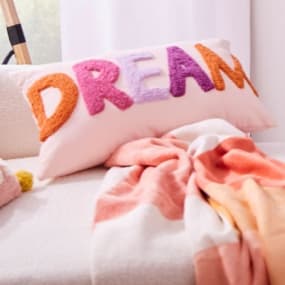 Dekoratives Kissen, auf dem das Wort "DREAM" in bunten Buchstaben zu sehen ist. Daneben liegt eine weiche, farbenfrohe Decke, die eine einladende und entspannte Atmosphäre schafft. Die sanften Farben und das verspielte Design verleihen dem Raum einen fröh