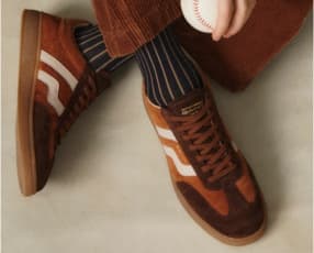 Ein Paar braune Retro Sneakers mit weißen Streifen, die aus einem weichen Material gefertigt sind. Die Schuhe sind mit gestreiften Socken kombiniert und passen gut zu einer lässigen Cordhose.