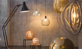 Ein stilvolles Arrangement von verschiedenen Lampen in einem modernen Raum. Eine große Stehlampe mit einem schwarzen Schirm steht links, während mehrere hängende Lampen in unterschiedlichen Formen und Materialien, darunter goldene und geflochtene Designs.