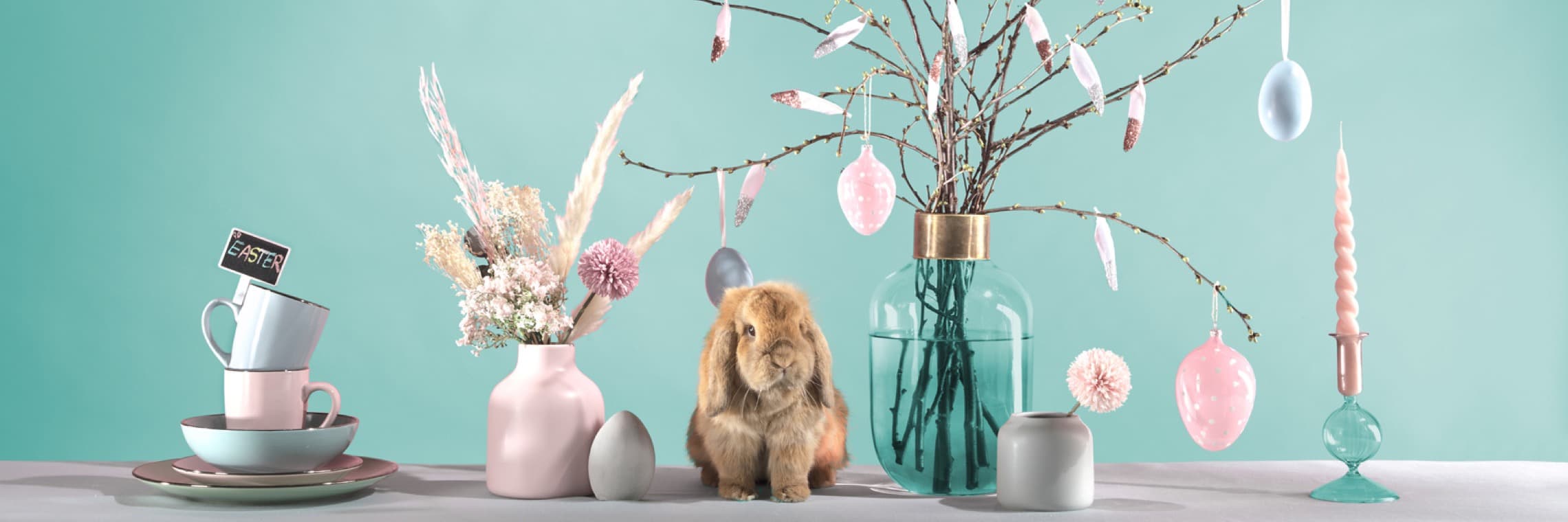 Ein niedliches Kaninchen sitzt vor einer festlich dekorierten Tischanordnung mit pastellfarbenen Tellern, Tassen, Vase mit Trockenblumen, verziertem Zweig mit bunten Ostereiern, Kerze