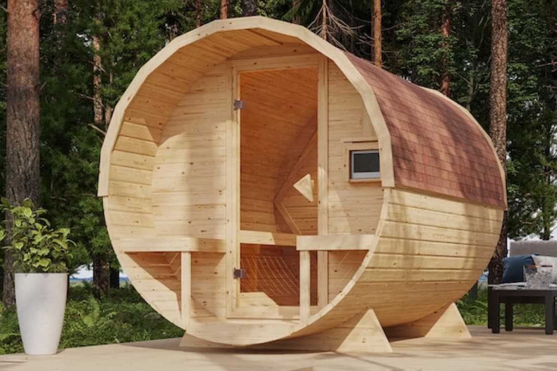 Runde Holzsauna im Freien, mit einer braunen Dachschindel und einer offenen Tür. Umgeben von Bäumen und einer weißen Pflanze im Topf.
