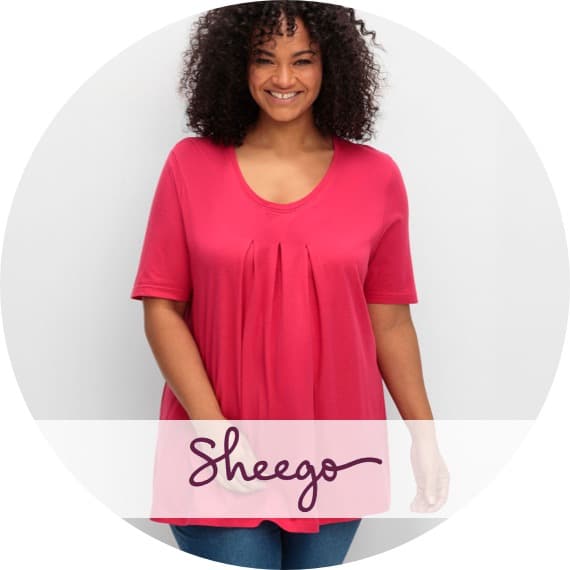 Sheego – Frau trägt locker fallendes pinkes T-Shirt mit Falten vorn und Jeans vor hellem Hintergrund